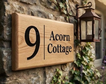 May include: A rectangular light brown wooden house sign featuring a large black number 9 on the left side and the text Acorn Cottage in black lettering on the right side, separated by a thin vertical black line, mounted on a stone wall.