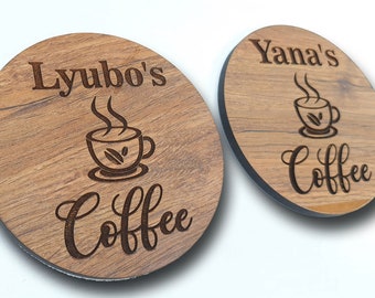 Op de afbeelding: Twee ronde houten onderzetters met een zwarte rand. De onderzetters zijn gegraveerd met de tekst "Lyubo's Coffee" en "Yana's Coffee" en een gestileerd beeld van een kopje koffie met stoom.