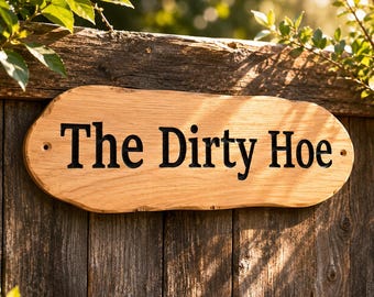 Op de afbeelding: Een ovaalvormig houten bord met een natuurlijke rand, voorzien van de tekst "The Dirty Hoe" gegraveerd in zwarte letters. Het bord bevat twee kleine voorgeboorde gaten aan de linker- en rechterkant voor montage op een verticaal oppervlak.