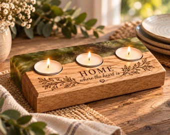 May include: A rectangular candle holder made of light brown wood and green resin, featuring three circular cutouts for tea light candles. The wood surface is engraved with the text "HOME where the heart is" and decorative leaf illustrations.