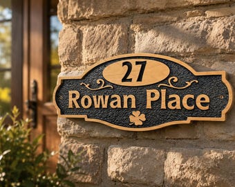 Op de afbeelding: Een ovaalvormig huisnummerbord met een lichte houten rand en een zwart getextureerd midden. Het bord bevat het nummer 27 bovenaan, de tekst Rowan Place in het midden en een klein klavertjevier-icoon onderaan, alles in licht hout.