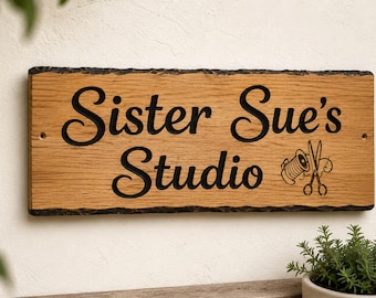 Op de afbeelding: Een rechthoekig houten bord met een natuurlijke rand, voorzien van de gegraveerde zwarte tekst "Sister Sue's Studio" en een afbeelding van een garenklosje en een schaar. Het bord heeft twee kleine voorgeboorde gaten aan de zijkanten.