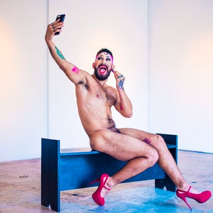 Puede incluir: Una persona con tacones rojos y maquillaje, sentada en un banco negro, tomando una selfie con un teléfono inteligente.