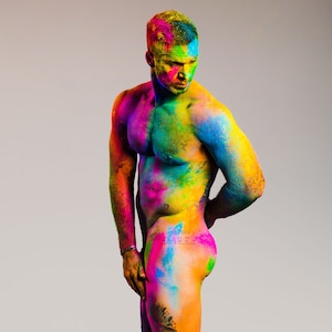 May include: A person covered in colorful powder, with a rainbow of colors on their skin and body. The person is standing with their arms at their sides, looking down.