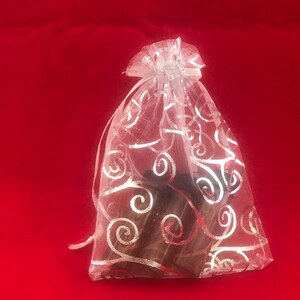Könnte beinhalten: Ein kleiner, durchsichtiger weißer Geschenkbeutel mit silbernem Wirbelmuster, gebunden mit einem Band. Der Beutel ist mit dunkel gefärbten Gegenständen gefüllt, vor einem leuchtend roten Hintergrund.