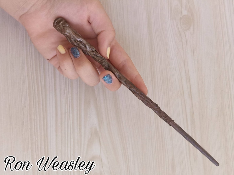 Puede incluir: Una varita marr&oacute;n con un mango texturizado y un extremo c&oacute;nico. La varita se sostiene en una mano con u&ntilde;as pintadas. El texto "Ron Weasley" est&aacute; escrito en una fuente de guion en la parte inferior de la imagen.
