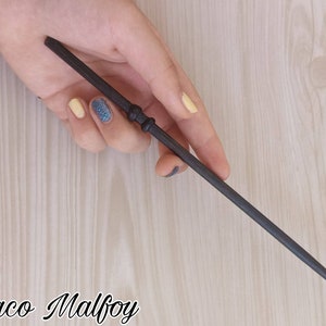 Puede incluir: Una varita gris oscuro con un mango detallado y un cuerpo delgado. La varita se sostiene en una mano con u&ntilde;as pintadas. El texto "Draco Malfoy" est&aacute; escrito en cursiva en la parte inferior de la imagen.