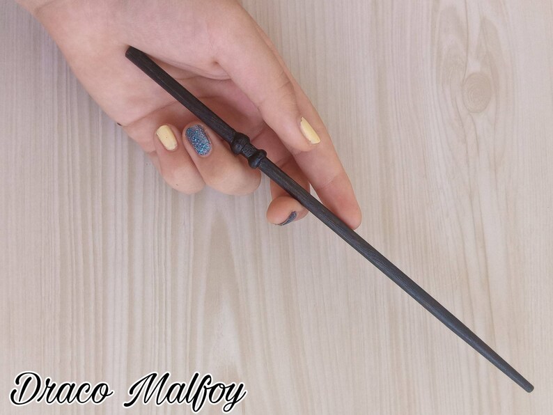 Puede incluir: Una varita gris oscuro con un mango detallado y un cuerpo delgado. La varita se sostiene en una mano con u&ntilde;as pintadas. El texto "Draco Malfoy" est&aacute; escrito en cursiva en la parte inferior de la imagen.