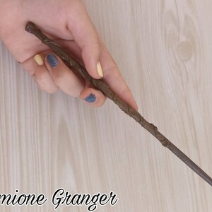 Puede incluir: Una varita marr&oacute;n con un mango texturizado y un extremo c&oacute;nico se sostiene en una mano. La varita est&aacute; etiquetada "Hermione Granger" en una fuente cursiva. El fondo es un patr&oacute;n de vetas de madera clara.