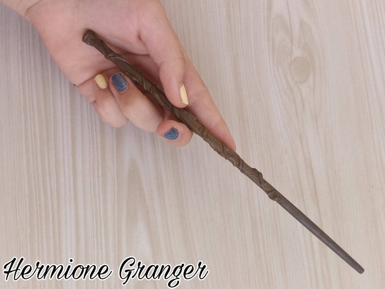 Puede incluir: Una varita marr&oacute;n con un mango texturizado y un extremo c&oacute;nico se sostiene en una mano. La varita est&aacute; etiquetada "Hermione Granger" en una fuente cursiva. El fondo es un patr&oacute;n de vetas de madera clara.