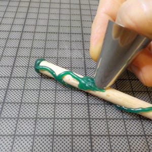 May include: A close-up of a wooden dowel being decorated with green icing. A metal piping tip is used to apply the icing in a decorative pattern. The dowel is on a gray grid surface.