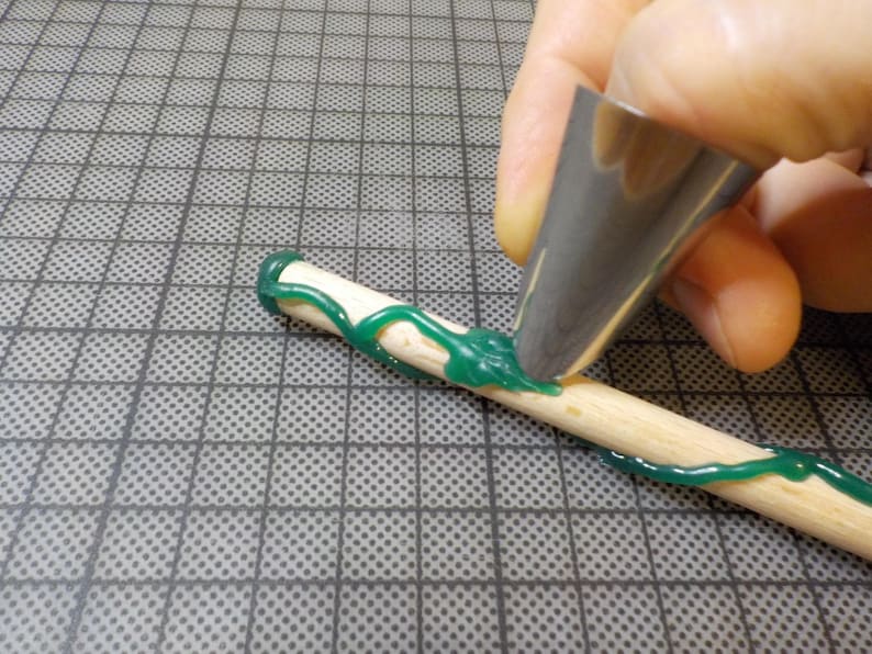 May include: A close-up of a wooden dowel being decorated with green icing. A metal piping tip is used to apply the icing in a decorative pattern. The dowel is on a gray grid surface.