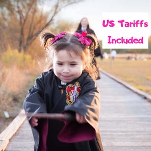 Könnte beinhalten: Ein Kind in einem schwarzen Umhang mit burgunderfarbenem Futter und Wappen, das einen Zauberstab hält. Das Kind befindet sich auf einem Holzsteg, im Hintergrund ist eine Person zu sehen. Der Text "US Tariffs Included" ist sichtbar.