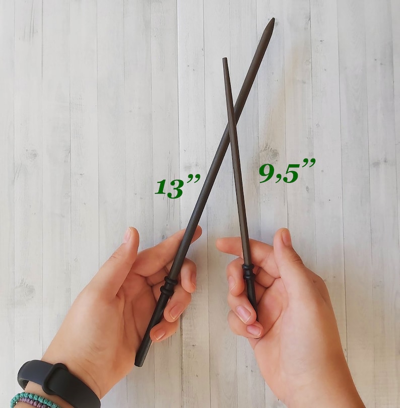 Puede incluir: Dos varitas de madera marr&oacute;n oscuro, una de 33 cm de largo y la otra de 24 cm, se cruzan sobre un fondo claro con vetas de madera. Las varitas tienen mangos detallados y puntas afiladas, lo que sugiere un tema m&aacute;gico.
