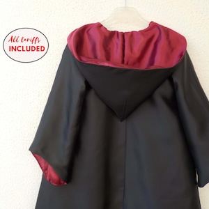 Peut inclure: Cape noire avec capuche doublée de satin bordeaux. La cape est suspendue à un cintre blanc contre un mur blanc. Le texte "All tariffs included" est dans un cercle blanc.