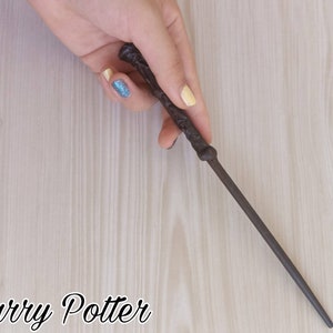 Puede incluir: Una varita marr&oacute;n oscuro con detalles intrincados, sostenida sobre una superficie clara de vetas de madera. La varita es delgada y parece ser de madera. El texto "Harry Potter" est&aacute; en la parte inferior de la imagen.