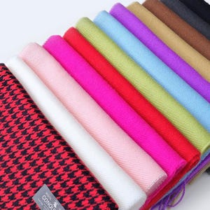 Puede incluir: Una colección de bufandas coloridas dispuestas en diagonal. Las bufandas muestran una variedad de colores lisos y estampados, incluido un diseño de pata de gallo rojo y negro con flecos. Una etiqueta con el texto "CONQUISTA" es visible en la bufanda roja.