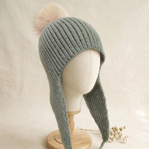 May include: A sage green ribbed knit hat featuring long ear flaps, braided ties ending in tassels, and a soft, light pink pom-pom on the crown. The hat is displayed on a neutral-coloured mannequin head stand.