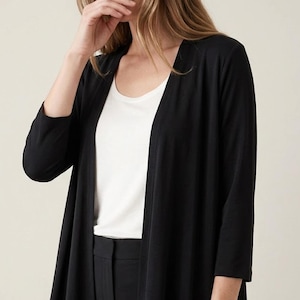 Puede incluir: Un cárdigan negro abierto por delante con mangas tres cuartos, usado sobre una camiseta sin mangas blanca de cuello redondo y combinado con pantalones negros. El cárdigan presenta una silueta fluida que cae por debajo de la cintura.