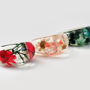 May include: Three clear resin bangles, each containing preserved flowers in red, orange, and green. The bangles are oval-shaped and rest on a white surface. The flowers are visible within the transparent resin, creating a unique and artistic look.