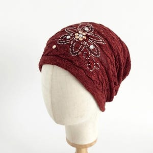 以下が含まれることがあります： ケーブル編みのデザインが施された、バーガンディ色のニット帽。 小さな白とベージュのキラキラ光るビーズで作られた花の模様が飾られています。 マネキンの頭に帽子が飾られています。