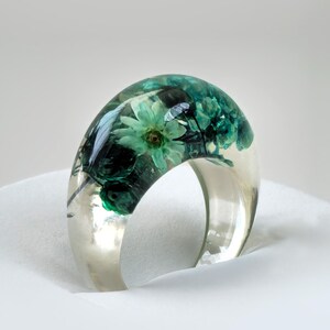 May include: A clear resin ring with embedded teal and green flowers. The ring has a wide band and a domed top, showcasing the preserved floral arrangement. The ring is set against a white background, highlighting its transparent quality.