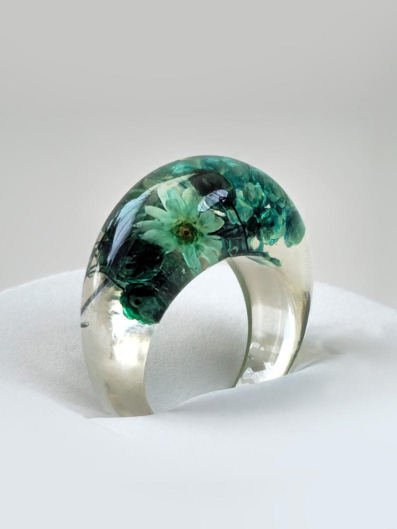 May include: A clear resin ring with embedded teal and green flowers. The ring has a wide band and a domed top, showcasing the preserved floral arrangement. The ring is set against a white background, highlighting its transparent quality.