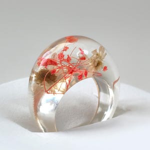 May include: A clear resin ring with embedded dried flowers in shades of red and beige. The ring has a rounded, dome-like top and a clear band. The ring is set against a white background.
