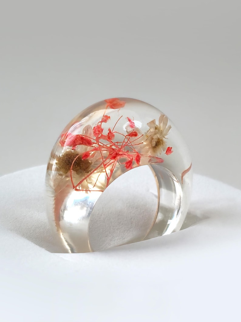 May include: A clear resin ring with embedded dried flowers in shades of red and beige. The ring has a rounded, dome-like top and a clear band. The ring is set against a white background.