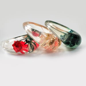 May include: Three clear resin bangles, each containing preserved flowers in red, peach, and green hues. The resin is transparent, showcasing the delicate floral details. The bangles are displayed on a white surface.