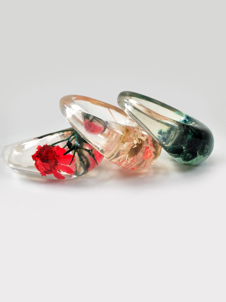 May include: Three clear resin bangles, each containing preserved flowers in red, peach, and green hues. The resin is transparent, showcasing the delicate floral details. The bangles are displayed on a white surface.