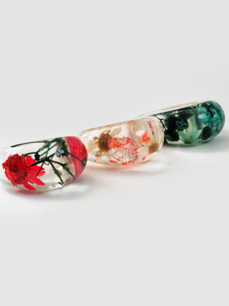 May include: Three clear resin bangles, each containing preserved flowers in red, orange, and green. The bangles are oval-shaped and rest on a white surface. The flowers are visible within the transparent resin, creating a unique and artistic look.