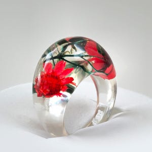 May include: A clear resin ring with a wide band, showcasing a vibrant red flower and green foliage encased within. The ring's transparent design allows the floral elements to be fully visible, creating a unique and artistic accessory.