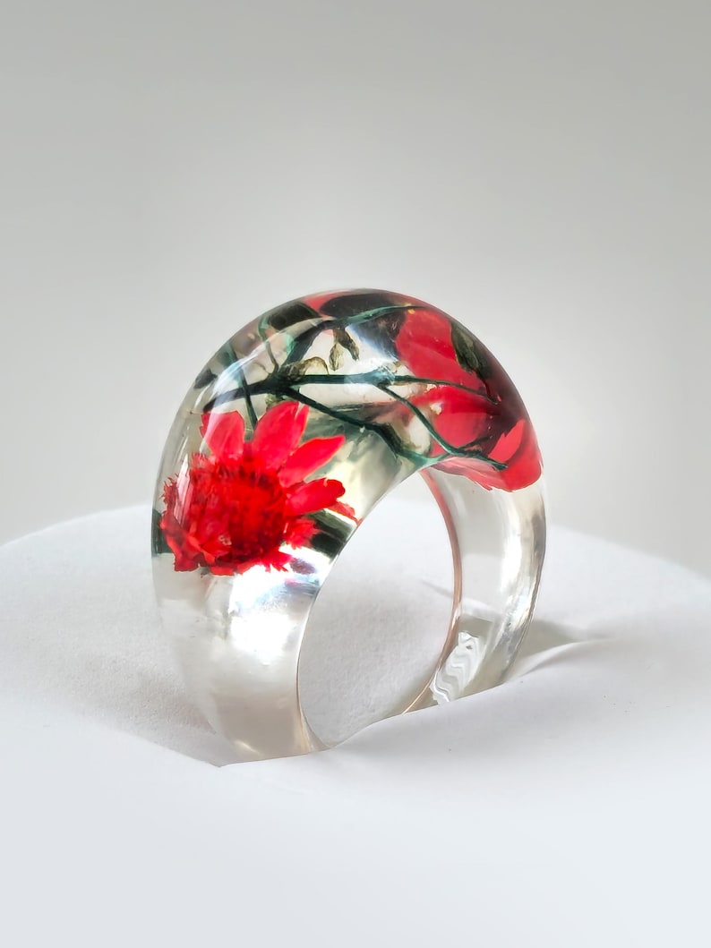May include: A clear resin ring with a wide band, showcasing a vibrant red flower and green foliage encased within. The ring's transparent design allows the floral elements to be fully visible, creating a unique and artistic accessory.
