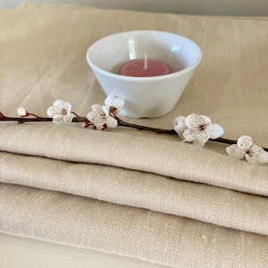 May include: A white ceramic bowl containing a pink candle, resting on a folded beige linen tablecloth with a sprig of white blossom.