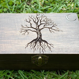 May include: A dark brown wooden box with a hinged lid and a metal latch. The top of the box features an engraved design of a tree with roots and a crescent moon.