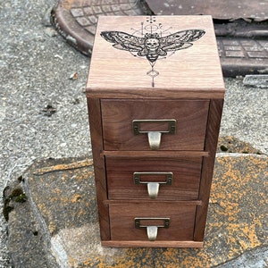 May include: A wooden drawer organizer with three drawers and a top panel featuring a black and white engraved design of a moth with geometric patterns.