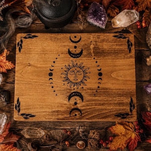 May include: A rectangular wooden board featuring a central black graphic of a sun with a face surrounded by moon phases. The four corners of the board are decorated with black leaf illustrations. The wood grain is visible across the surface.