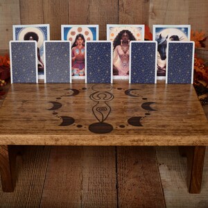 May include: A wooden tarot card reading table with a carved design of a woman surrounded by crescent moons. The table has four legs and is holding several tarot cards. The cards have a blue starry design on the back. The table is surrounded by fall foliage.