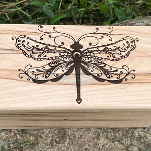 May include: A wooden box with a hinged lid. The lid is decorated with a laser-engraved design of a dragonfly with intricate details. The box is made of light wood with a natural grain.
