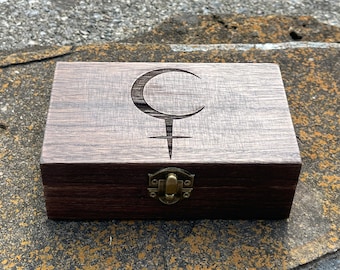 May include: A dark wood box with a carved crescent moon and cross design on the lid. The box has a brass latch.