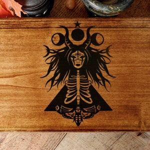 May include: A wooden box with a black and white design of a woman with horns, a skeleton, and a moth. The design is centered on the box and is surrounded by a dark brown wood grain.