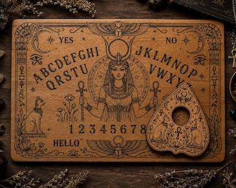 Könnte beinhalten: Ein rechteckiges Spirit-Board aus Holz mit einer gravierten Abbildung einer ägyptischen Gottheit, dem Alphabet, den Zahlen 1-8 sowie den Wörtern YES, NO und HELLO. Eine passende herzförmige Planchette mit einem Loch liegt auf dem Brett.