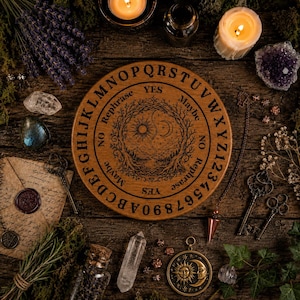 May include: A round wooden spirit board featuring an engraved sun and moon design at the center, surrounded by the alphabet, numbers zero through nine, and the words Yes, No, Maybe, and Rephrase, arranged in a circular pattern on the board.