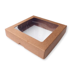 Può includere: Una scatola di cartone marrone con una finestra di plastica trasparente nella parte superiore. La scatola è vuota e ha una leggera ombra sullo sfondo bianco.