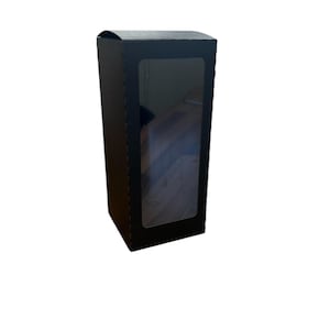 Puede incluir: Una caja de cartón negra con una ventana transparente en la parte delantera. La caja está diseñada para contener un producto, como una vela o jabón.