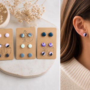 K&ouml;nnte beinhalten: Drei Sets von Ohrsteckern auf braunen Karten. Jede Karte zeigt zwei Paar Ohrringe in verschiedenen Farben und Mustern, darunter marmorierte und unifarbene Designs. Ein Ohrring wird am Ohr einer Person gezeigt, mit einem schwarz-wei&szlig;en Marmormuster.