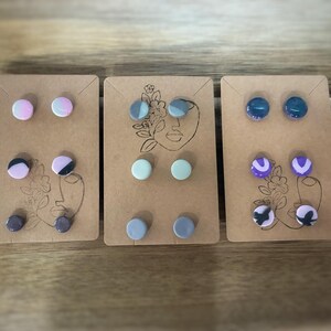 K&ouml;nnte beinhalten: Drei Sets handgefertigter Ohrstecker. Jedes Set hat zwei Paar runde Ohrringe in verschiedenen Farben und Mustern, darunter Rosa, Blau, Lila und Schwarz. Die Ohrringe sind auf kleinen, braunen Karten mit einem minimalistischen Gesichtsdesign befestigt.