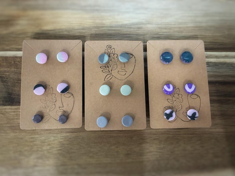 K&ouml;nnte beinhalten: Drei Sets handgefertigter Ohrstecker. Jedes Set hat zwei Paar runde Ohrringe in verschiedenen Farben und Mustern, darunter Rosa, Blau, Lila und Schwarz. Die Ohrringe sind auf kleinen, braunen Karten mit einem minimalistischen Gesichtsdesign befestigt.