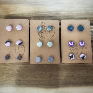 K&ouml;nnte beinhalten: Drei Sets von Ohrsteckern auf braunen Karten. Jede Karte zeigt eine Strichzeichnung eines Gesichts und florale Akzente. Die Ohrringe sind in verschiedenen Farben erh&auml;ltlich, darunter Rosa, Schwarz, Grau, Blau und Lila. Die Ohrringe sind rund.